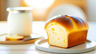 Une brioche dorée et légère au yaourt, tranchée et servie sur une assiette, à côté d'un pot de yaourt.