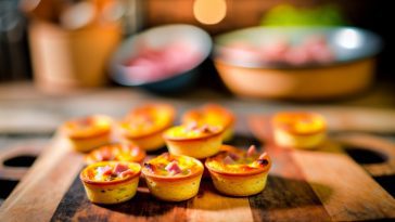 Des mini quiches lorraine sans pâte, disposées sur une planche en bois rustique, avec un fond de cuisine flou.
